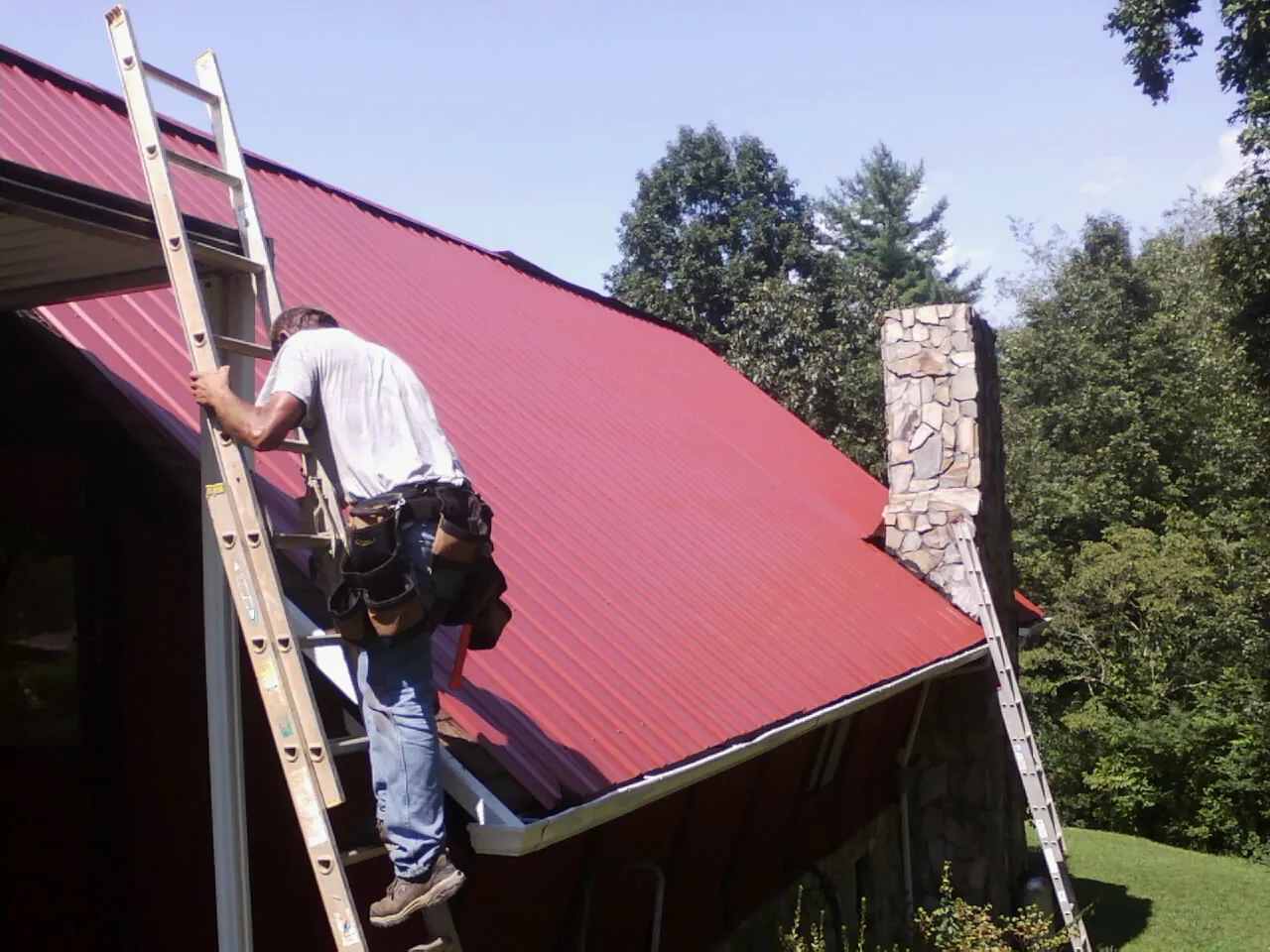 Roofer on a ladder beside a red metal roof and stone chimney — Soffit & Fascia Repair by Roofing Brothers in Preemption